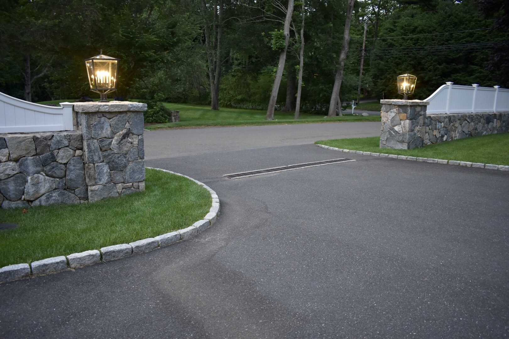 This image shows a driveway flanked by stone pillars with lit lanterns, a white fence, and trees in the background during the evening.