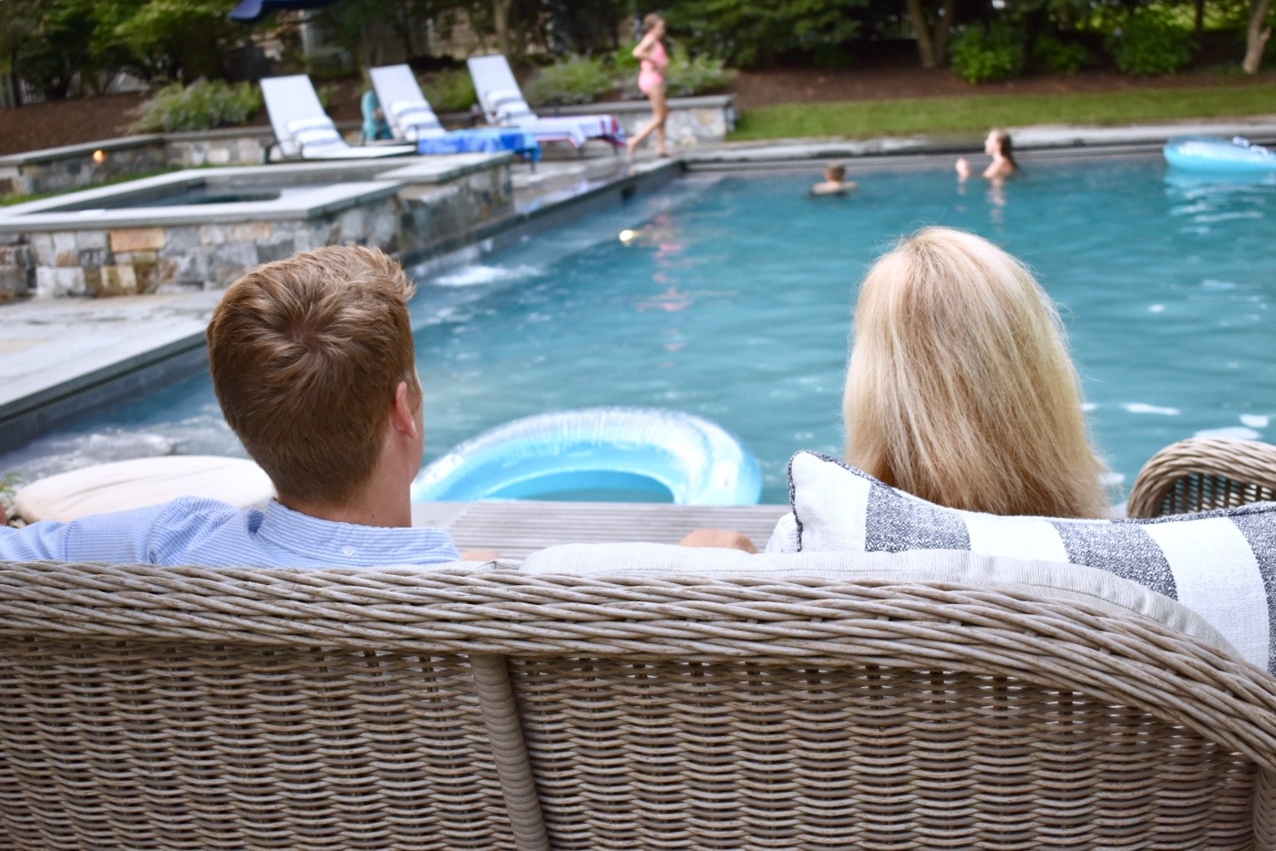 Two people are sitting on a wicker sofa by a pool, watching others swim on a sunny day with greenery in the background.