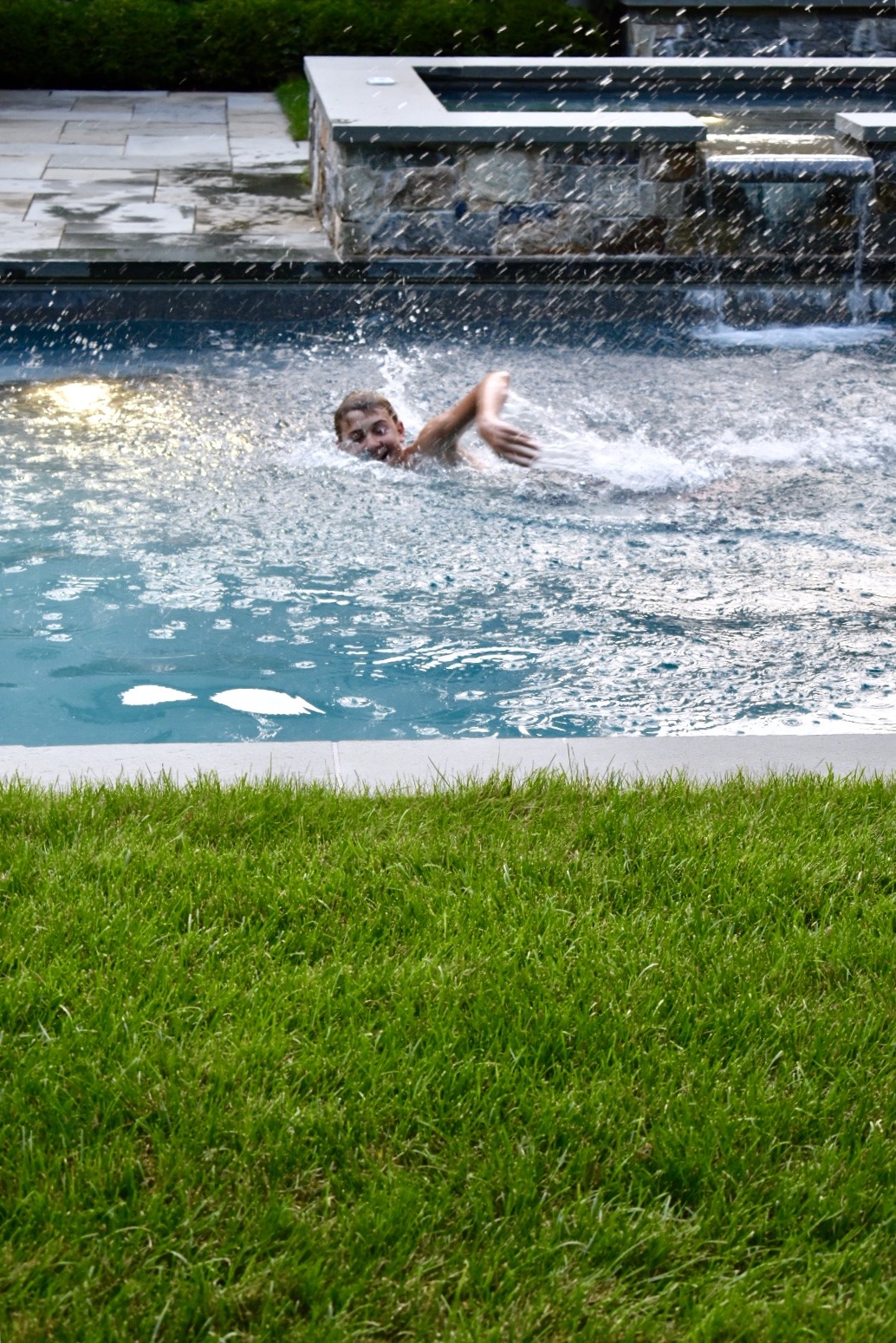 A person splashes into a pool, creating a dynamic water disturbance, against a backdrop of green grass and a water feature with cascading steps.
