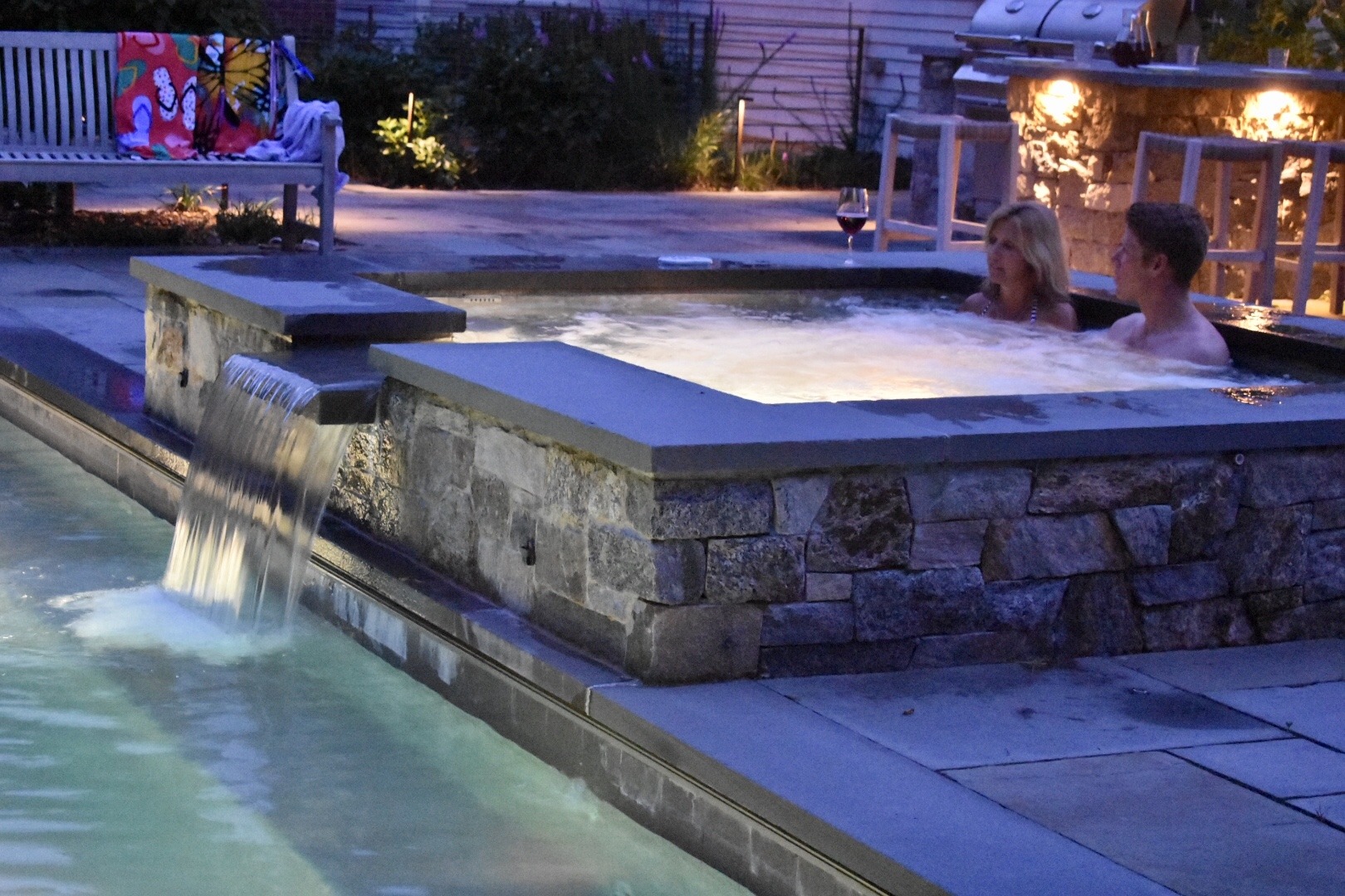 Two people relax in a hot tub with bubbling water at dusk. Nearby, water cascades from a stone feature into a serene pool.