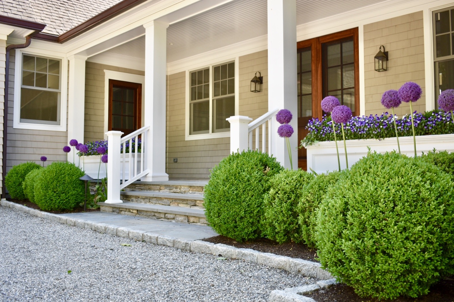 A charming home entrance with a covered porch, white columns, green bushes, purple flowers, and a gravel path under a clear blue sky.