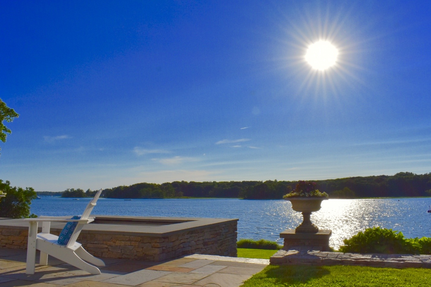 The image shows a tranquil lakeside view with an Adirondack chair on a patio, a stone planter, glistening water, and a bright sun in a clear blue sky.