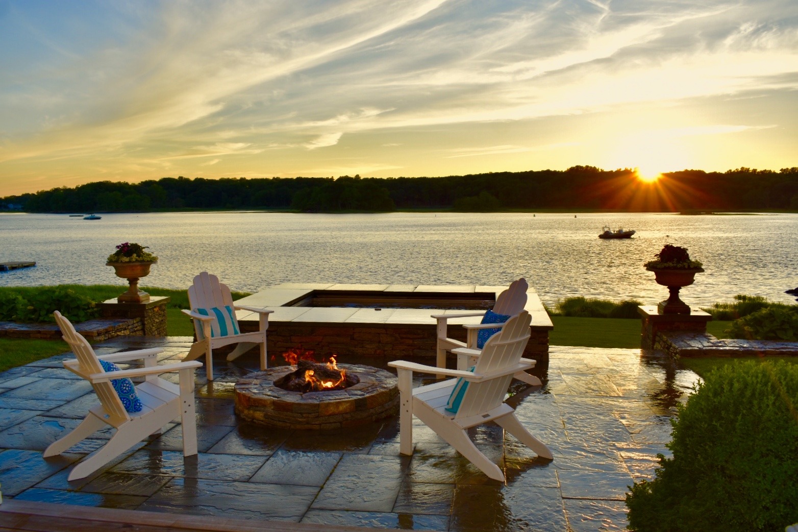 Two Adirondack chairs overlook a serene lake with a fire pit at sunset, with boats on the water and lush greenery surrounding the peaceful scene.