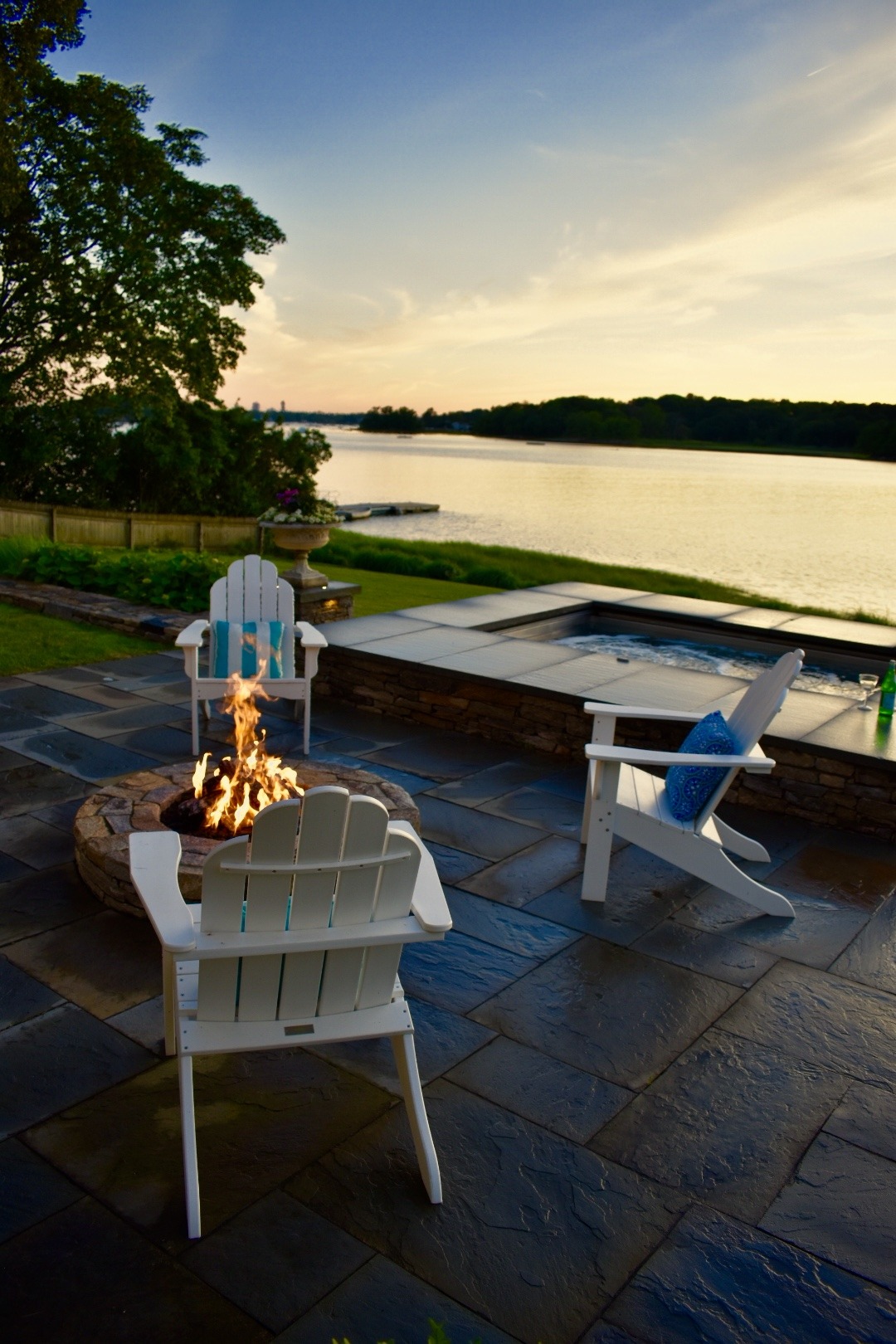 A serene outdoor setting with Adirondack chairs, a fire pit, overlooking a calm river during sunset, creating a tranquil and inviting atmosphere.