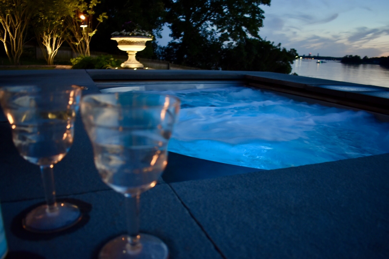 Two wine glasses sit on a surface next to a hot tub with glowing blue water. Evening setting, trees and a river visible in the background.