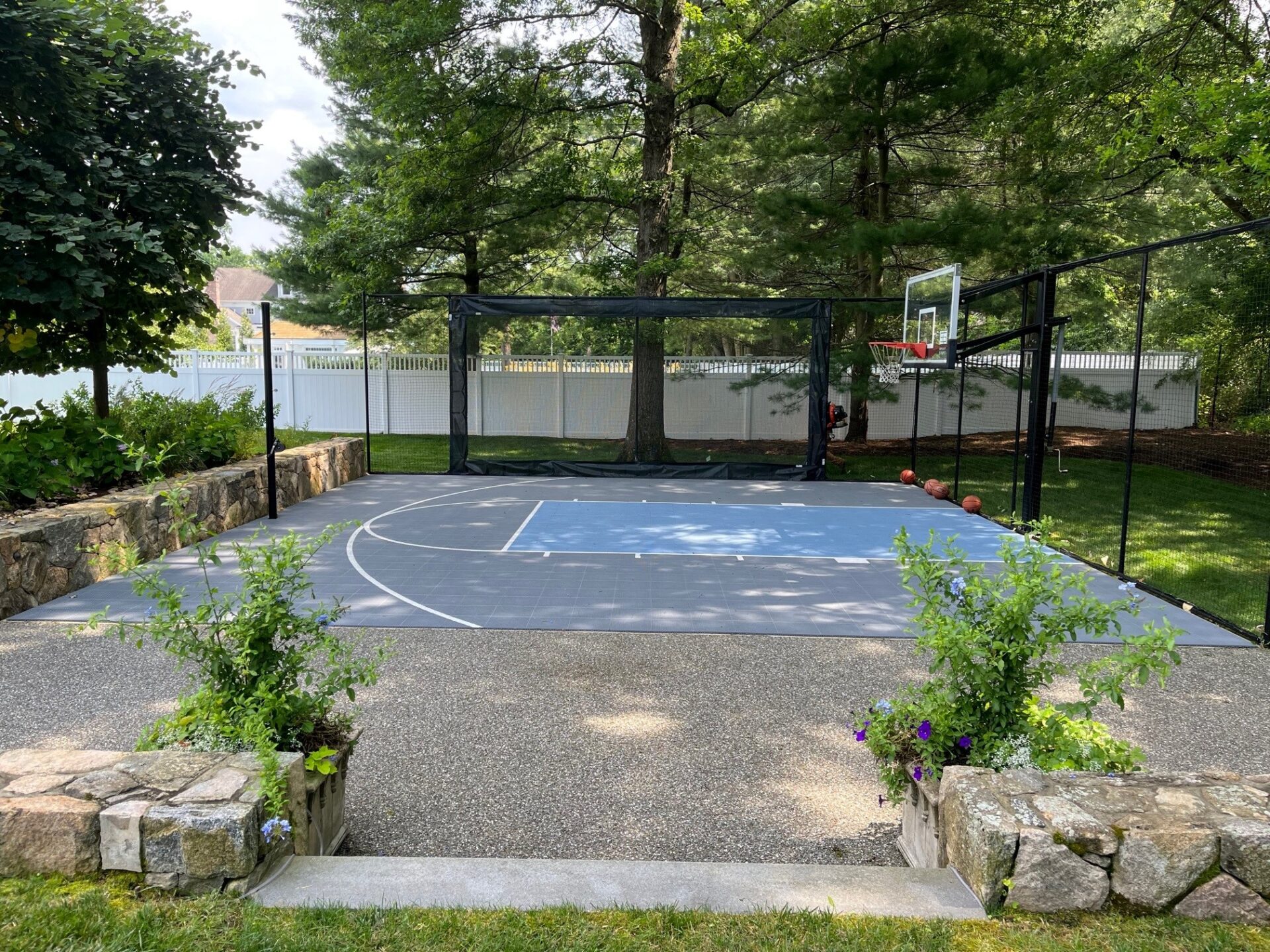 This image shows an outdoor basketball court with a fence, a basketball hoop, and several basketballs. There's greenery and a clear sky overhead.