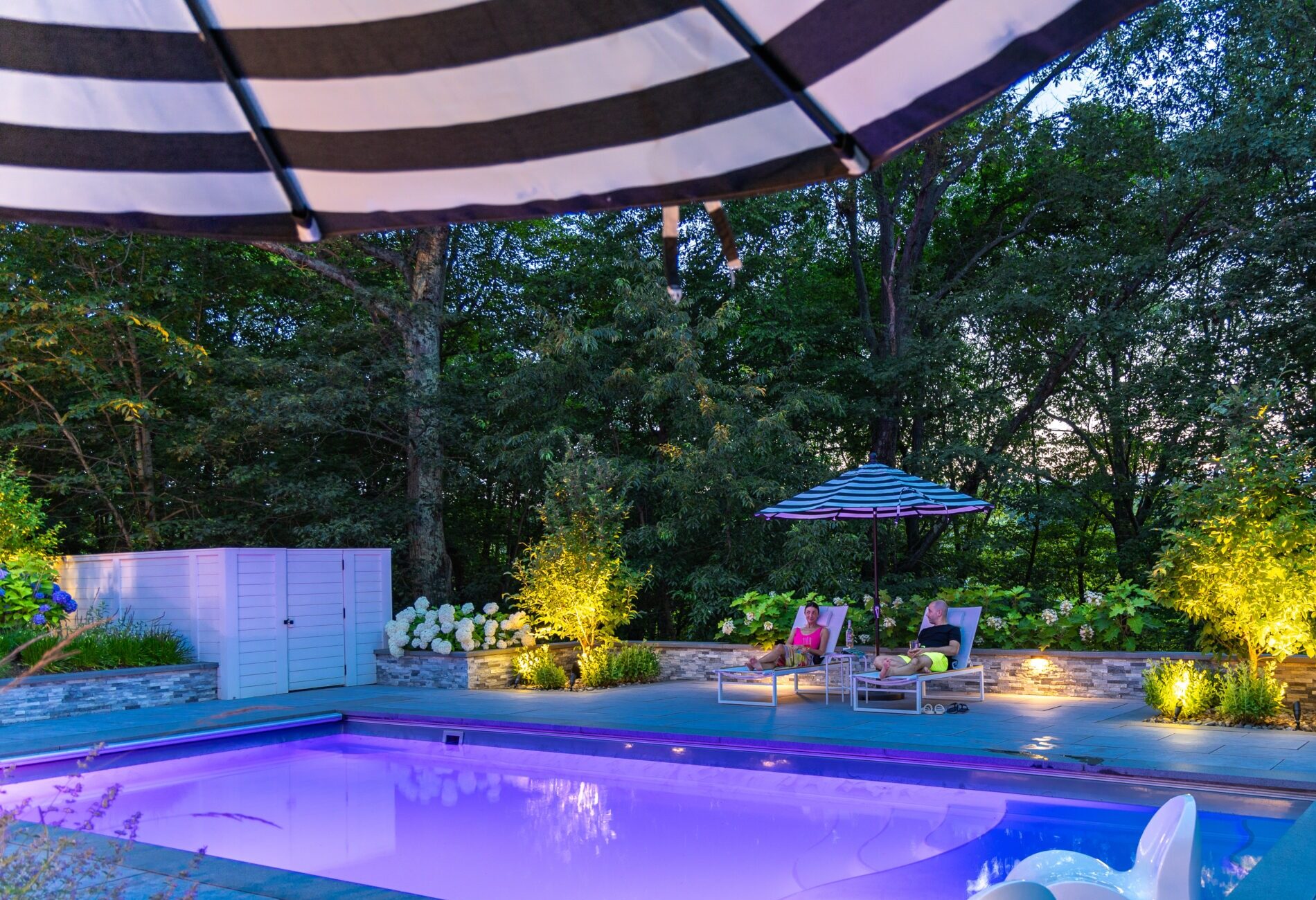 An outdoor pool area at dusk, with two people relaxing on lounge chairs under a striped umbrella, surrounded by trees and illuminated plants.