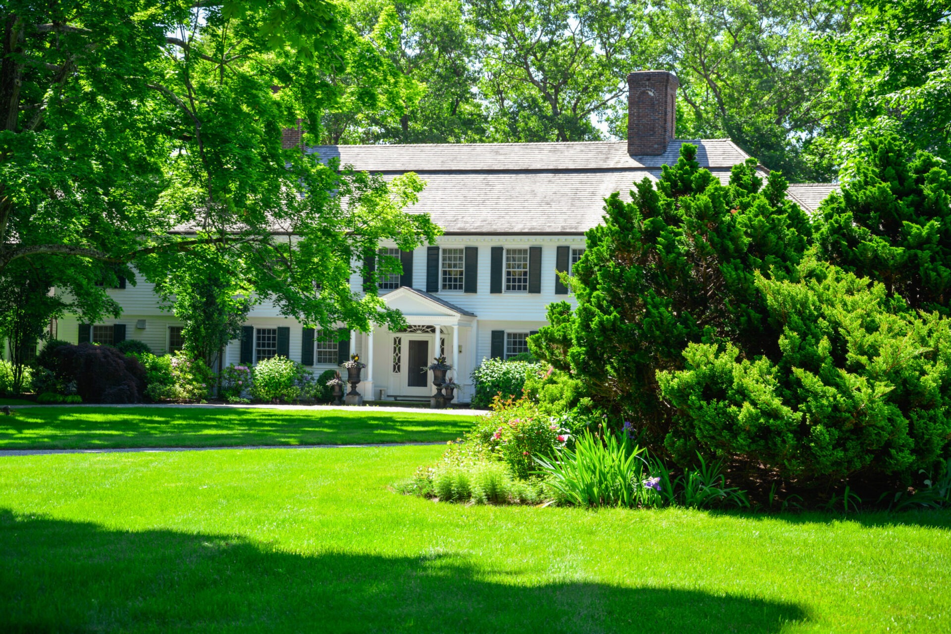 This is a two-story, white colonial house with green shutters surrounded by lush greenery, manicured lawn, and well-maintained garden beds on a sunny day.