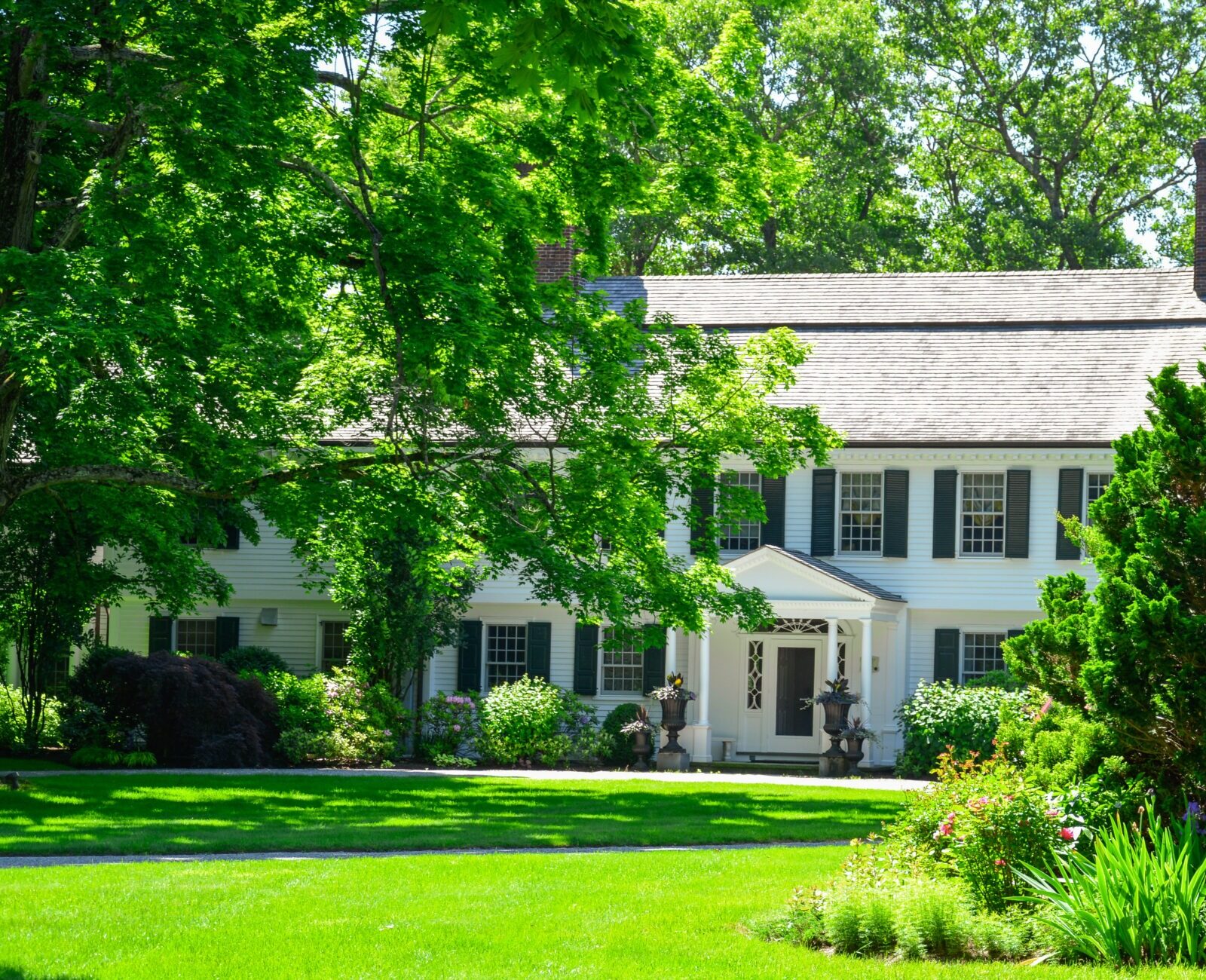 This is a two-story, white colonial house with green shutters surrounded by lush greenery, manicured lawn, and well-maintained garden beds on a sunny day.