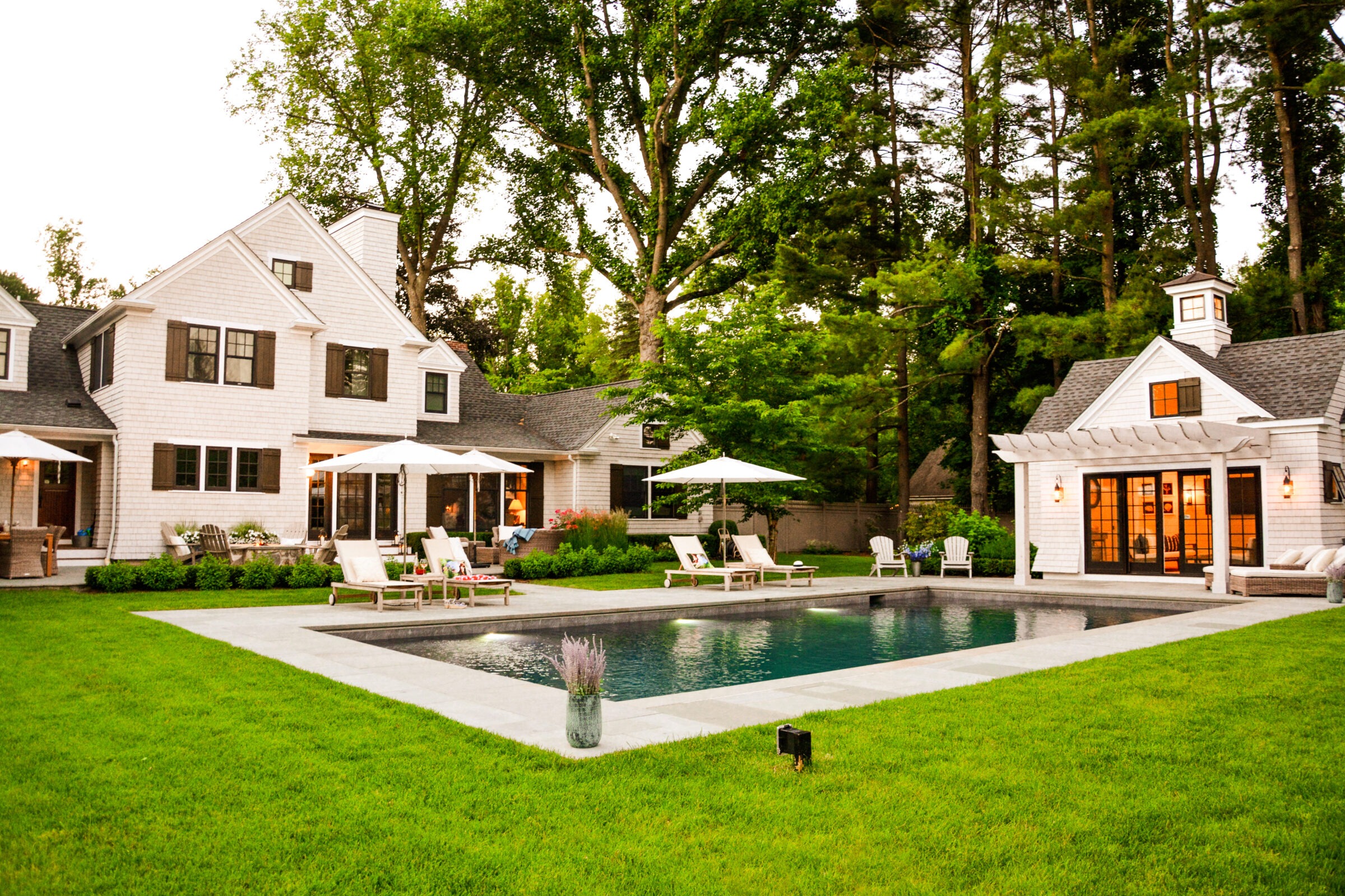 A spacious backyard with a rectangular swimming pool, loungers, and umbrellas, adjacent to an elegant, large white house surrounded by lush greenery.