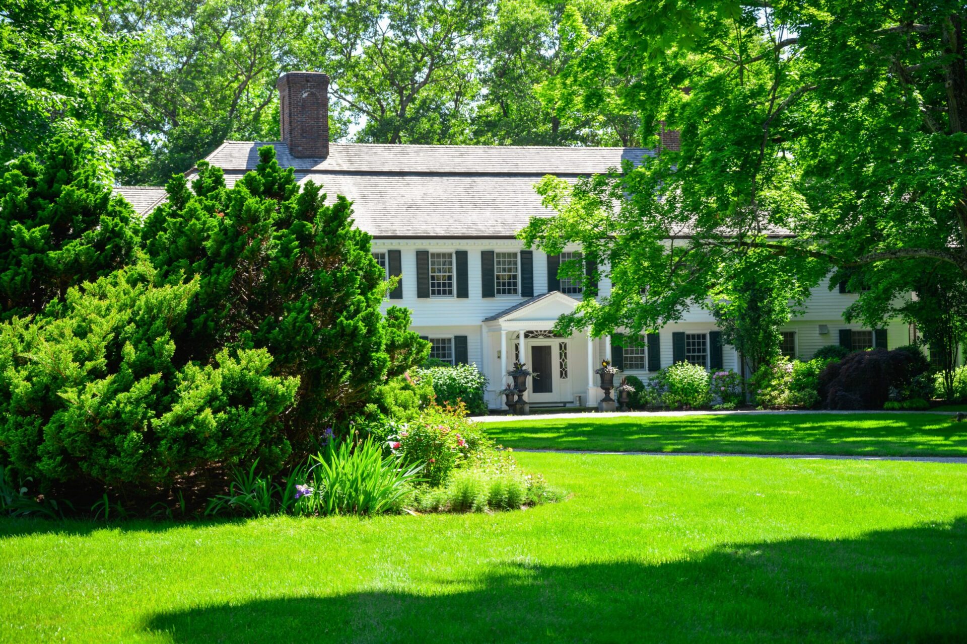 A two-story white house with black shutters, surrounded by lush greenery, under a clear blue sky. A well-manicured lawn enhances the tranquil setting.
