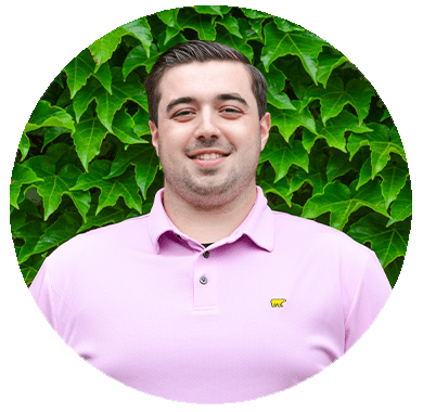 Portrait of a smiling person wearing a pink polo shirt with a logo, standing in front of a lush green ivy background. Image is circularly cropped.