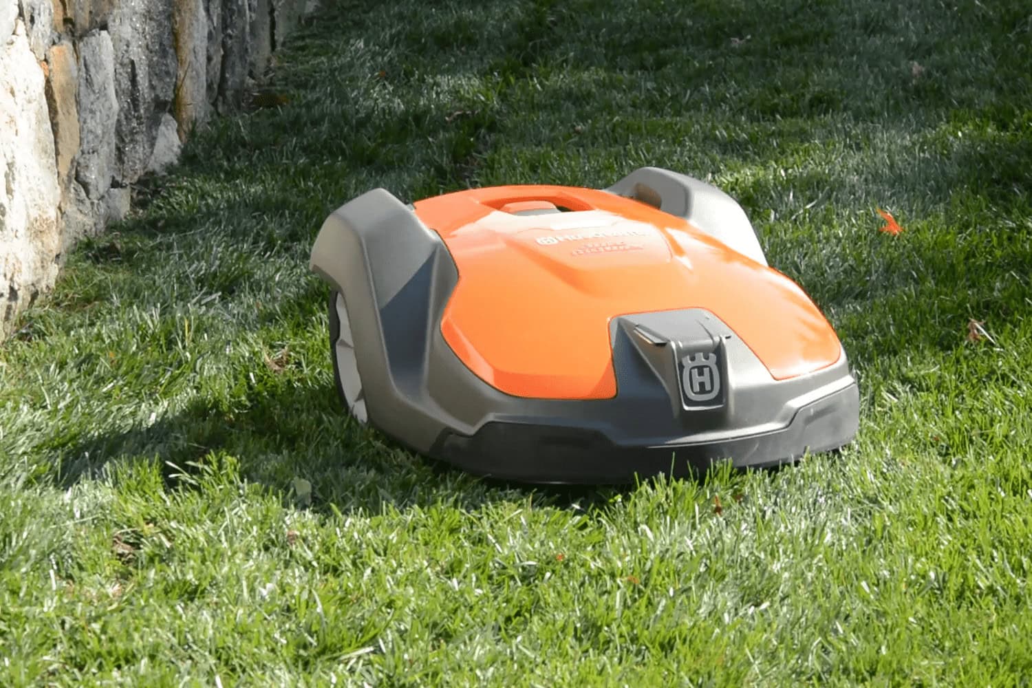 An orange and gray robotic lawn mower rests on a lush green lawn near a stone boundary under bright sunlight. The grass appears freshly trimmed.