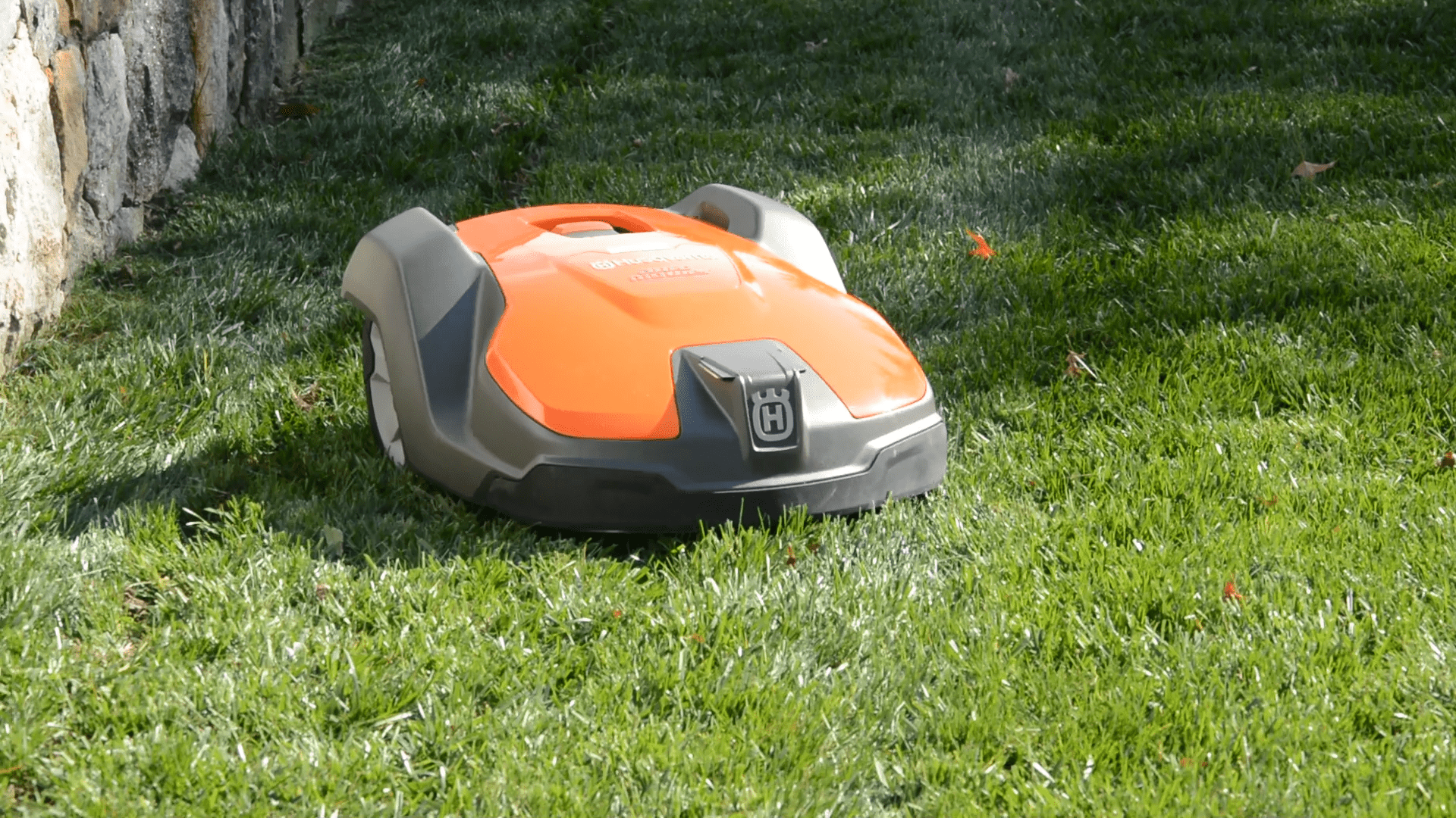 An orange and gray robotic lawn mower rests on a lush green lawn near a stone boundary under bright sunlight. The grass appears freshly trimmed.
