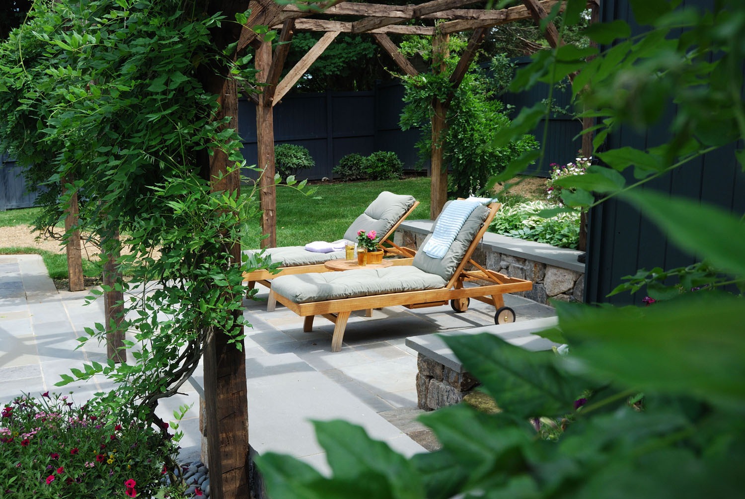 A serene garden with a wooden pergola, two reclining chairs with cushions, and a table with drinks surrounded by vibrant greenery and flowers.