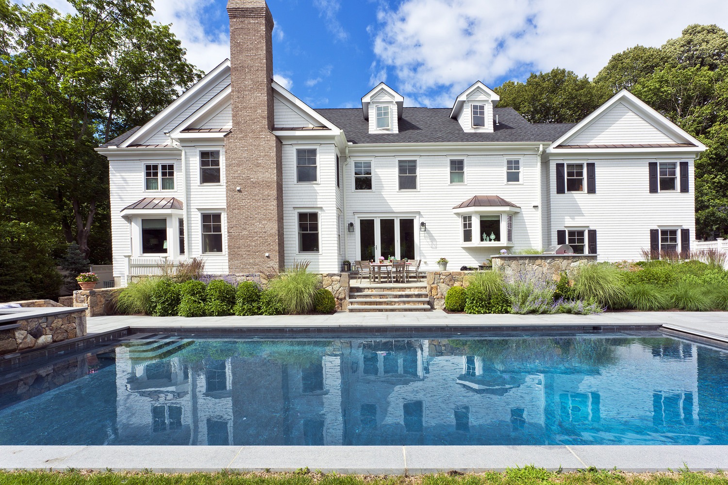 A large white house with multiple windows and chimneys reflects in a clear blue swimming pool surrounded by manicured landscaping under a sunny sky.