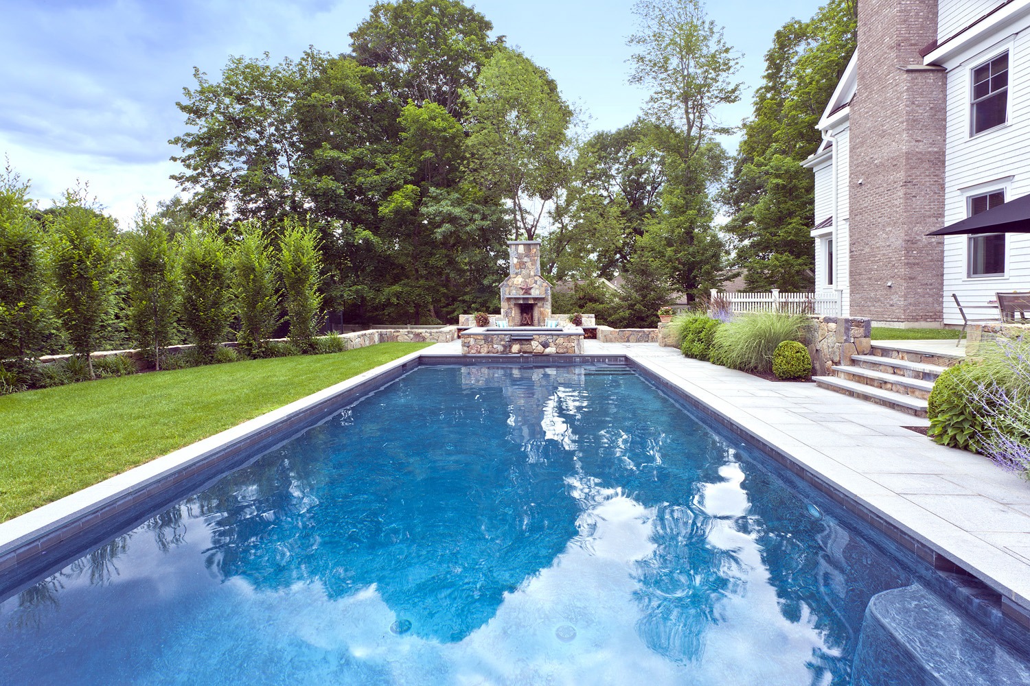 A serene backyard with a rectangular pool, stone fountain, lush greenery, and a brick house with white trim under a partly cloudy sky.