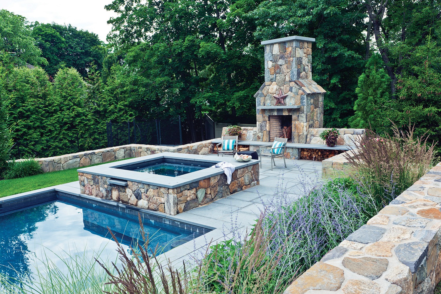 An outdoor space featuring a rectangular swimming pool, an integrated hot tub, a stone fireplace, patio chairs, surrounded by lush greenery and fencing.