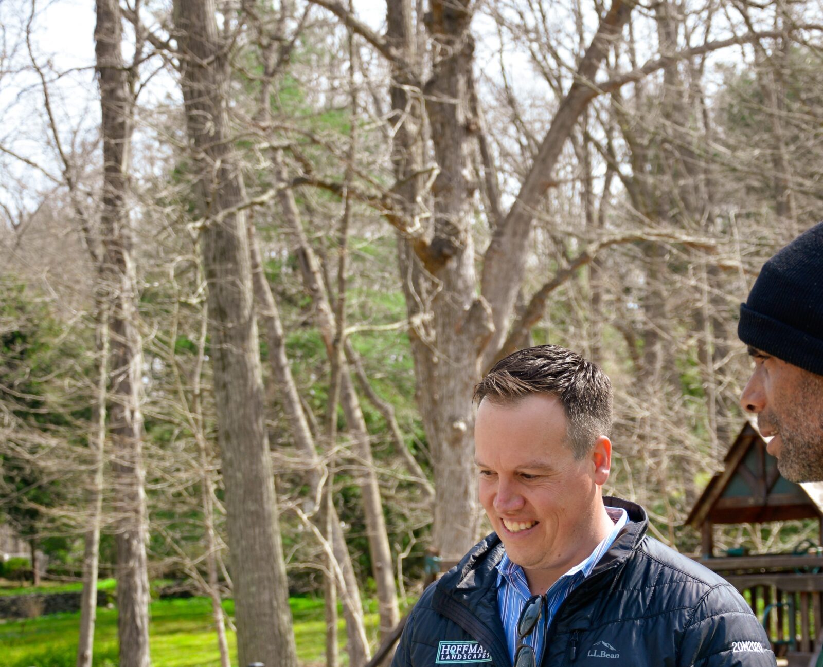 Two people are having a conversation outdoors. One person is gesturing, and both are wearing jackets with company logos. Trees and a fence surround them.