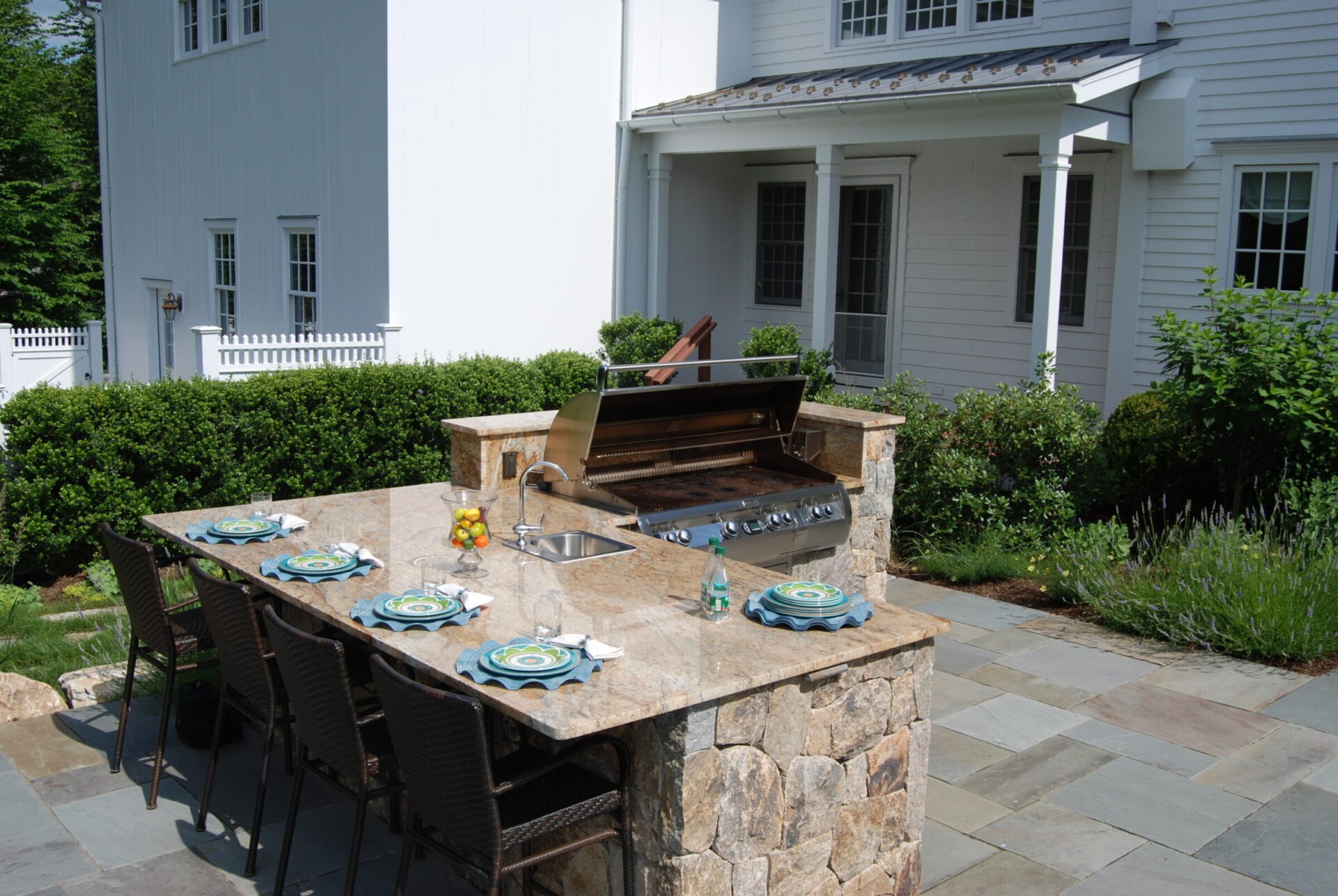 An outdoor kitchen with a grill, sink, and dining area set against a white house, adorned with stone countertops and surrounded by greenery.