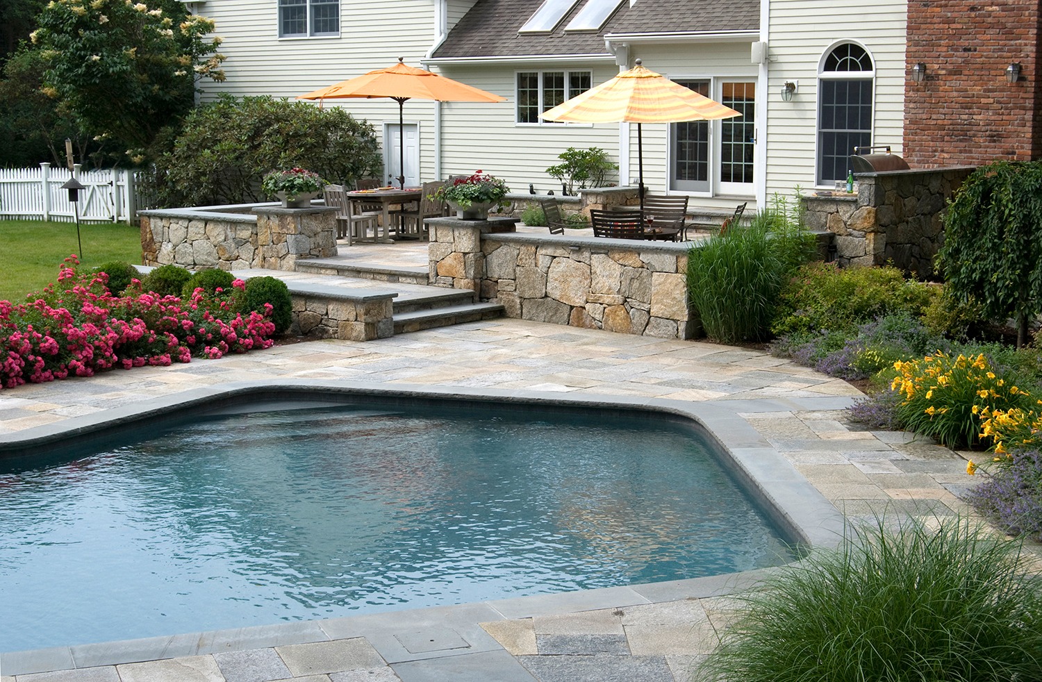 An elegant backyard featuring a swimming pool with stone coping, surrounded by lush gardens, patio furniture under umbrellas, adjacent to a residential home.
