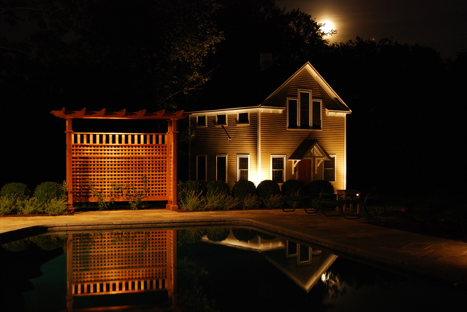 A night scene featuring a two-story house illuminated by warm light, a swimming pool in the foreground reflecting a pergola, and a rising moon.