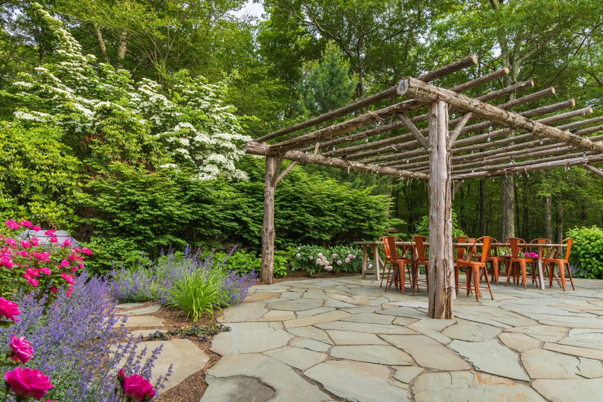 A tranquil garden patio with a wooden pergola, stone flooring, surrounded by lush greenery and colorful flowers, with a set of wooden chairs and a table.