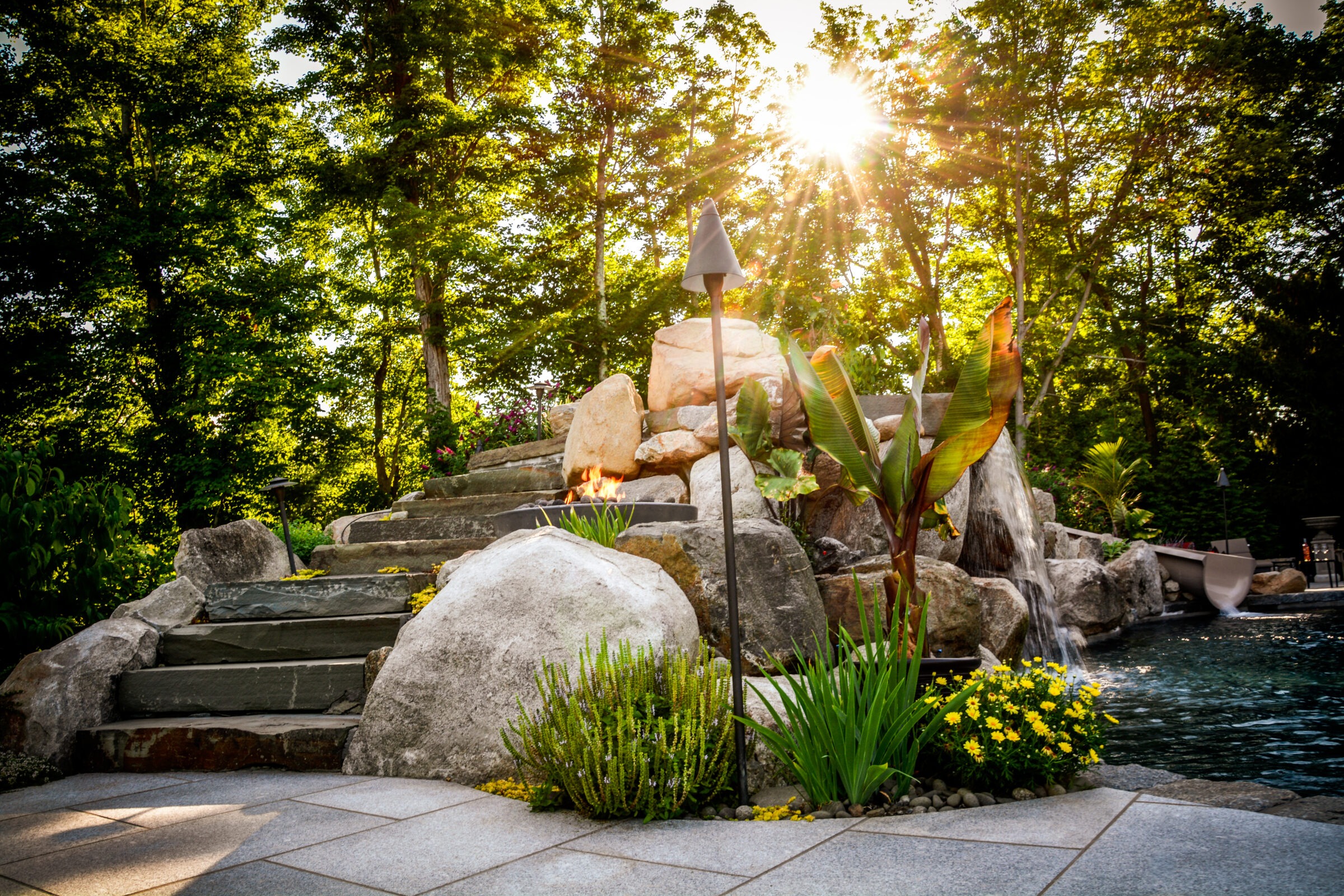 An elaborate garden with a stone waterfall, lush plants, and a sunburst through the trees, showcasing a tranquil, naturalistic landscape design.