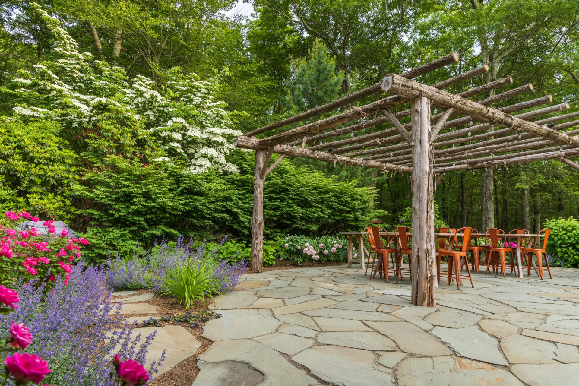 A serene garden patio with a wooden pergola, slate flooring, and blooming flowers. There's a table with chairs set up for outdoor dining.