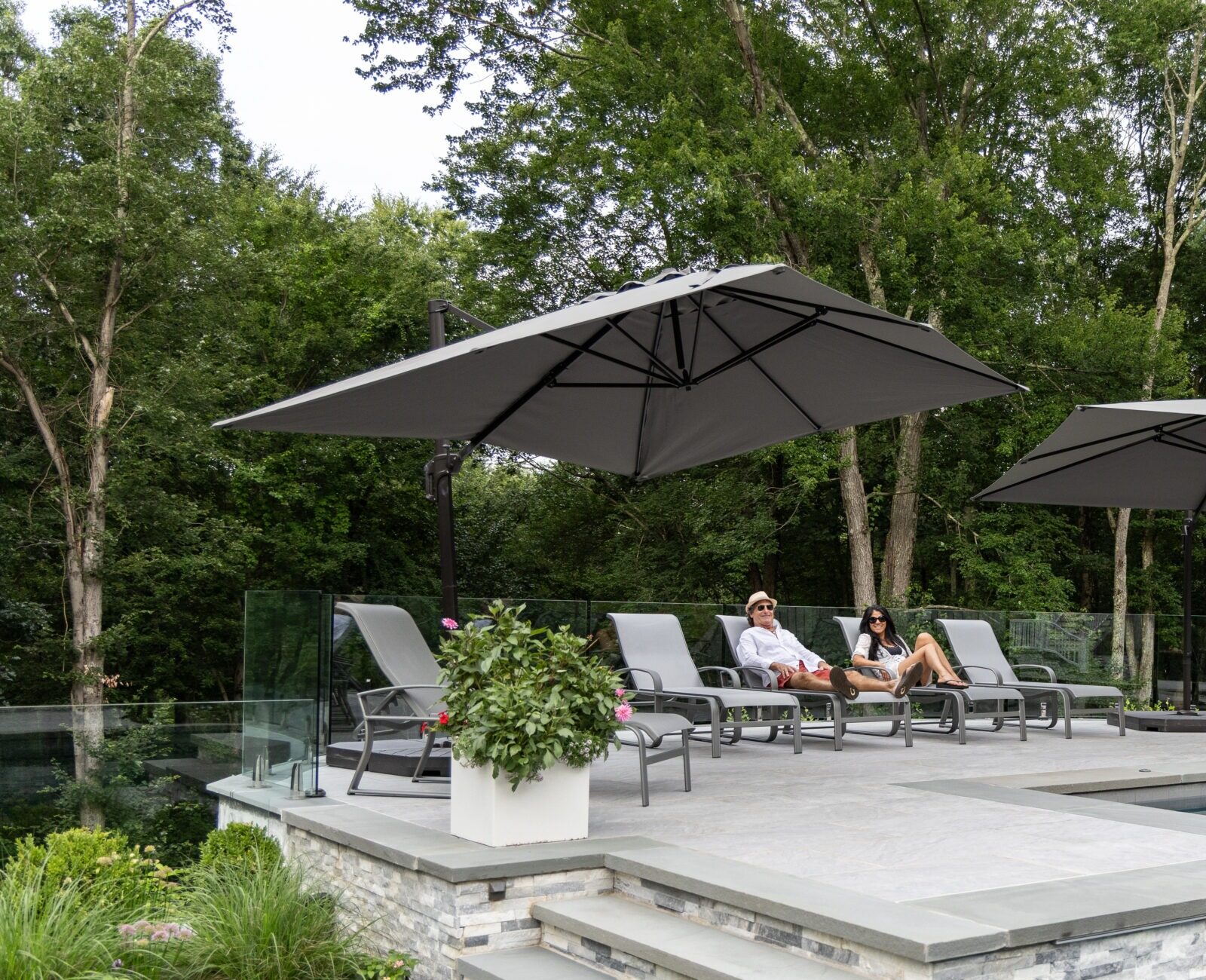 An outdoor patio with a swimming pool, loungers, and umbrellas. Two people are relaxing. Green trees surround the serene, landscaped area.