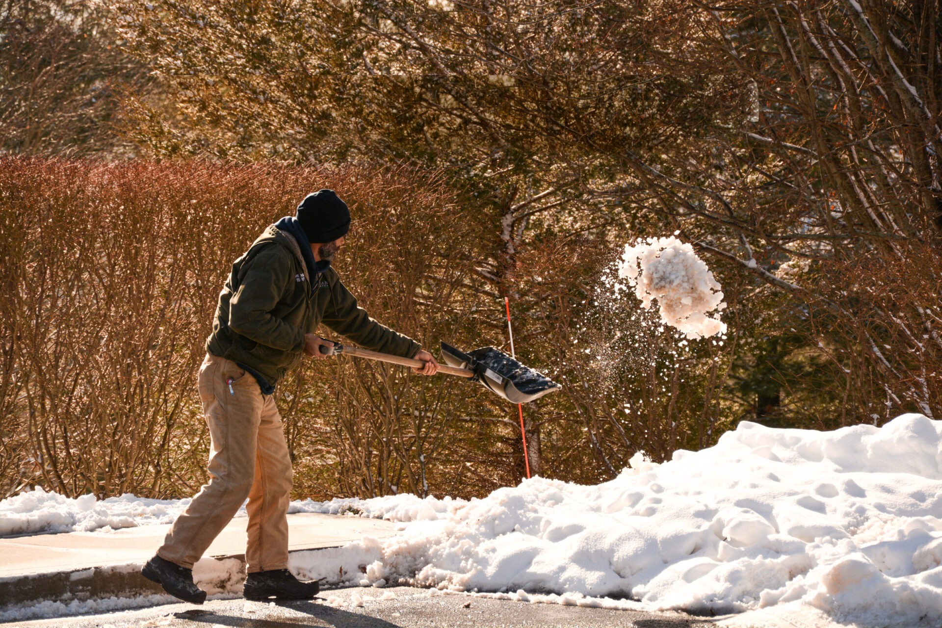 A person is throwing a snowball with a shovel mid-swing, capturing the snow in motion. They are bundled up against a wintry, sunny background.