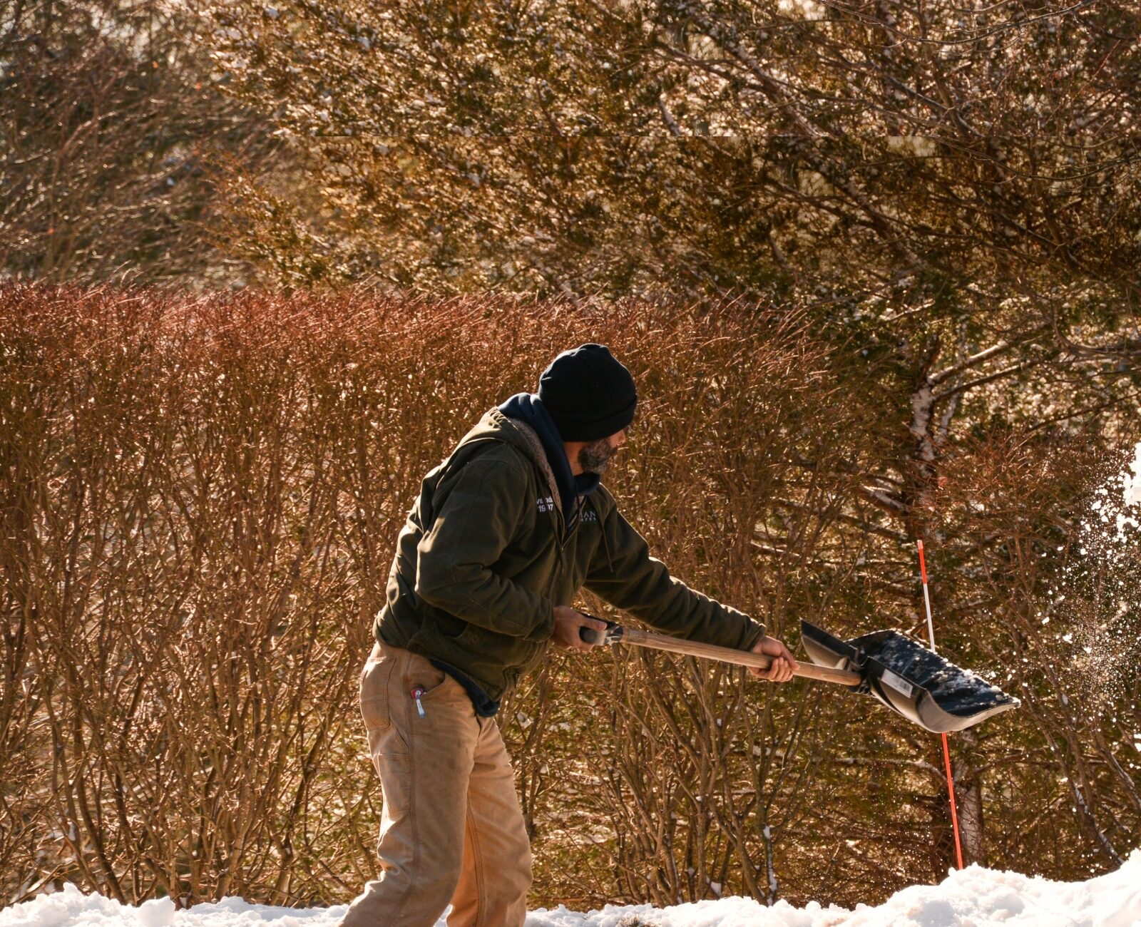 A person is throwing a snowball with a shovel mid-swing, capturing the snow in motion. They are bundled up against a wintry, sunny background.