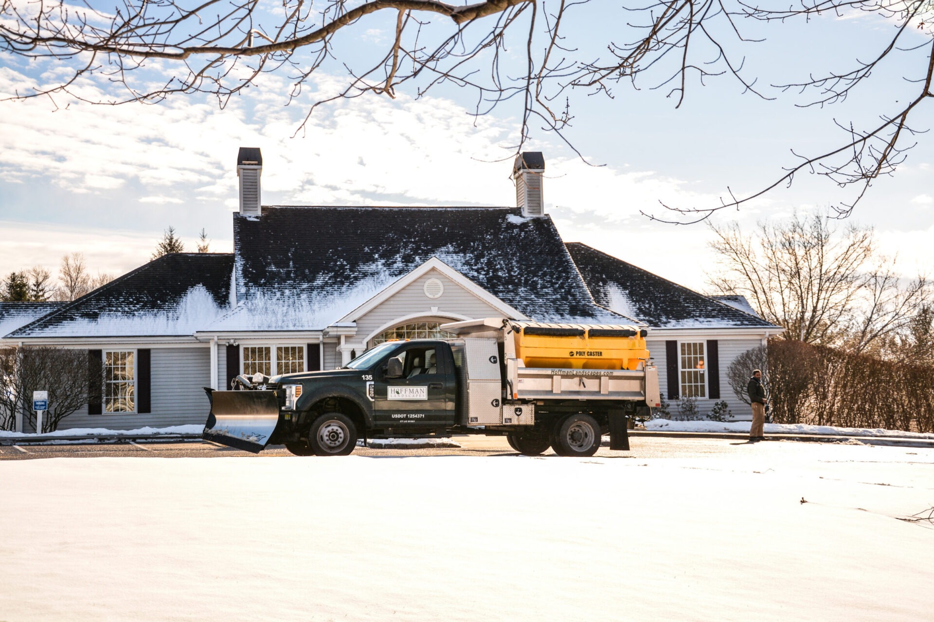 A snow-covered house is visible with a snow plow truck in the driveway. A person stands nearby on a clear, sunny winter day.