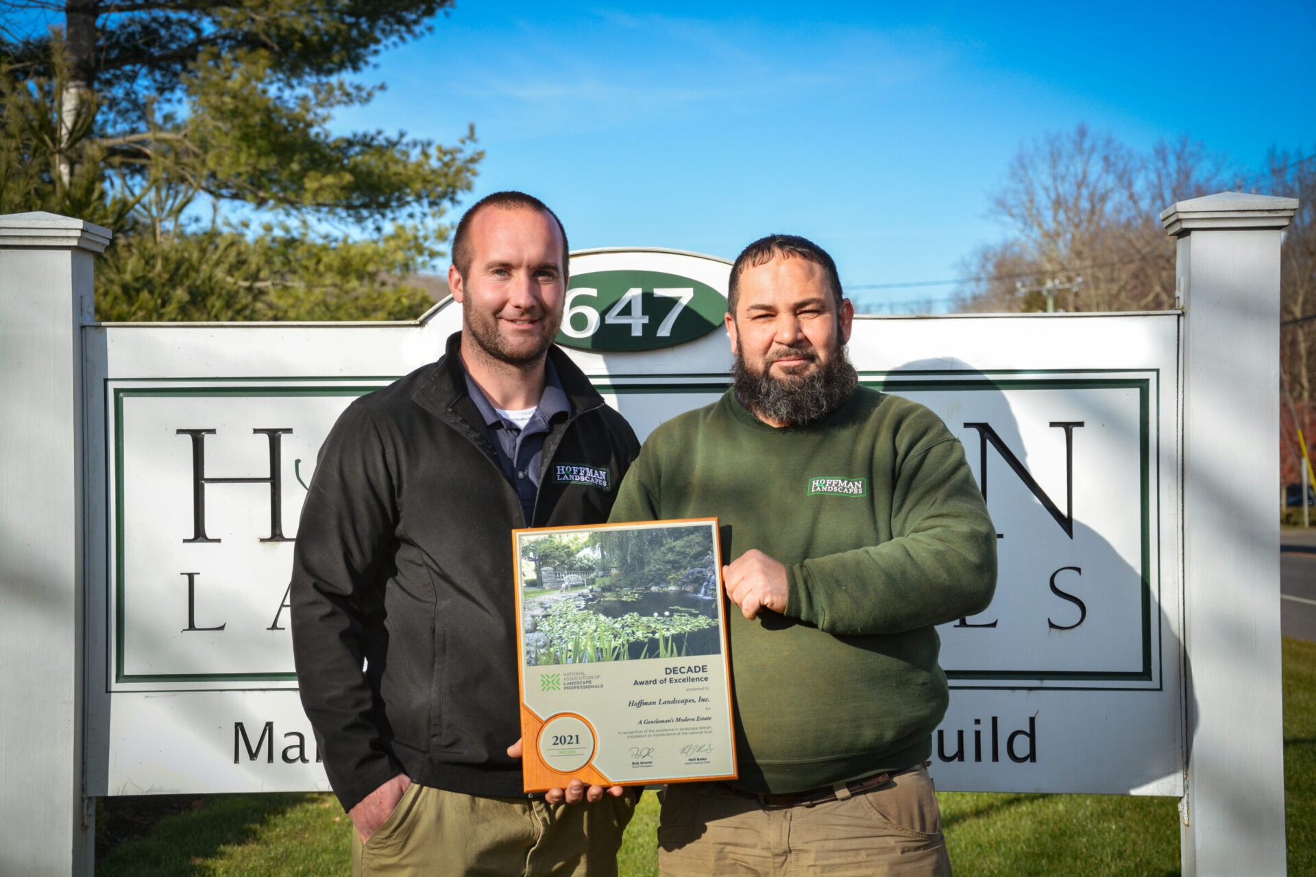 Two persons are standing in front of a sign labeled "Hoffman Landscapes," holding an award for excellence dated 2021. It is a sunny day.