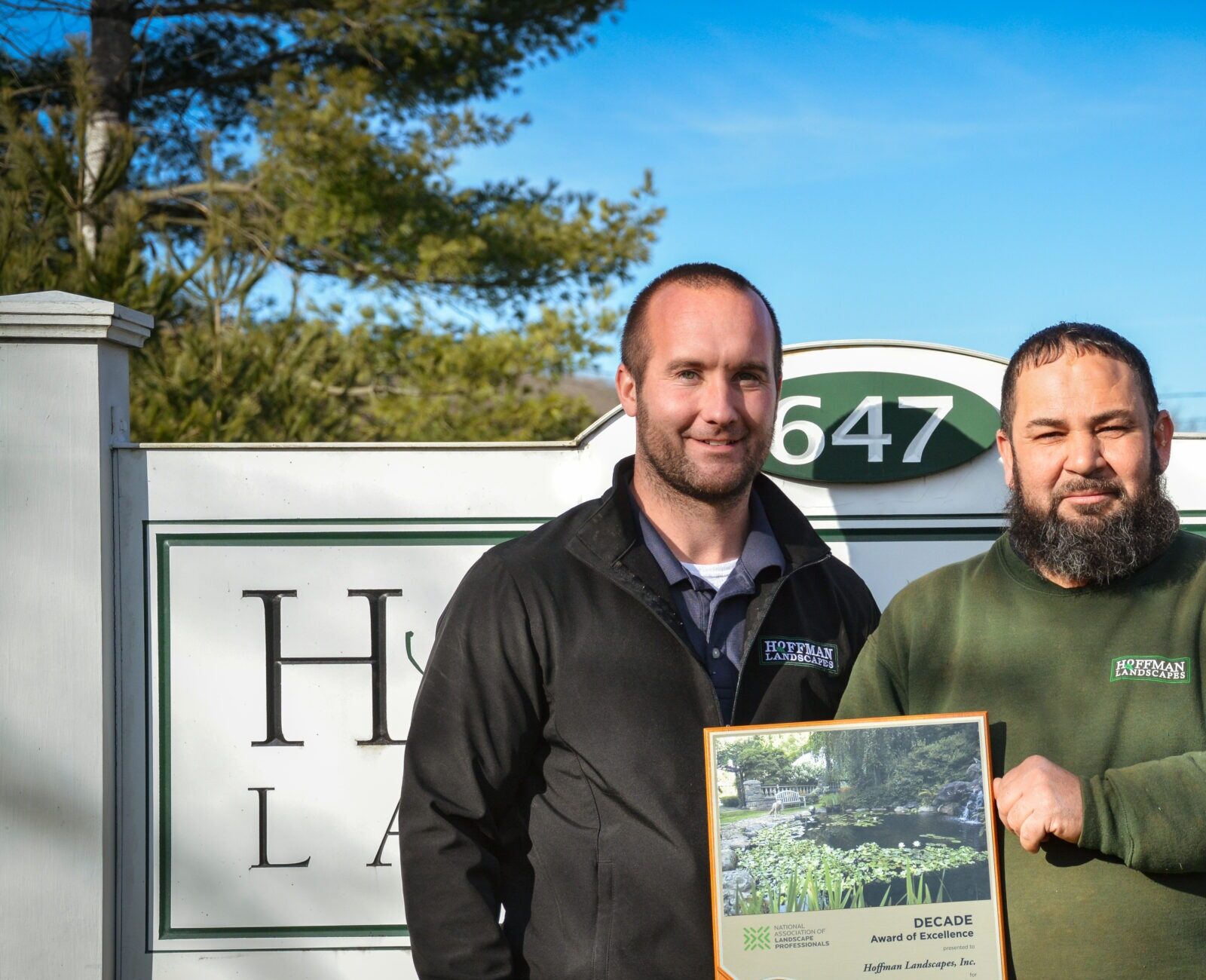 Two persons are standing in front of a sign labeled "Hoffman Landscapes," holding an award for excellence dated 2021. It is a sunny day.