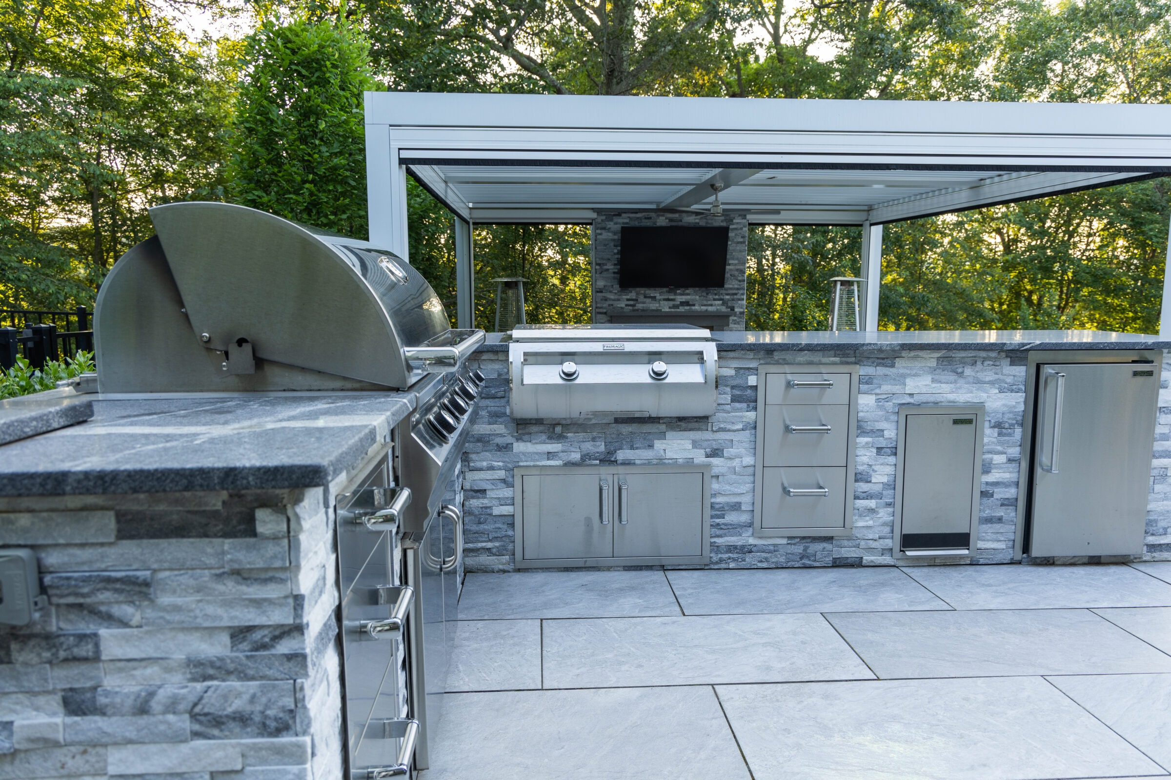 This image shows an outdoor kitchen with a stainless steel grill, sink, refrigerator, and a mounted television, set against a backdrop of greenery.