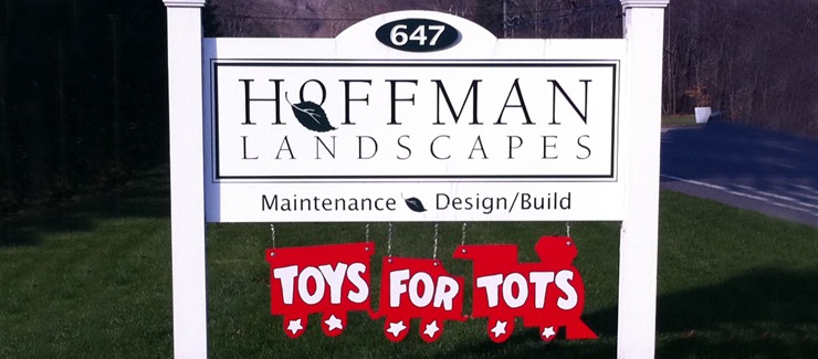 The image shows a sign for "Hoffman Landscapes" with an additional "Toys for Tots" banner hanging below it, indicating support for a charity.