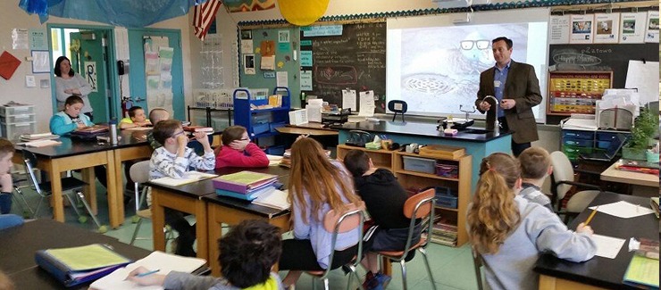 A person stands at the front of a classroom, gesturing towards a projection screen, with attentive children seated at desks, and a person watching from the back.