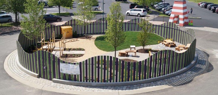 The image shows a circular outdoor play area enclosed by a patterned fence, featuring trees, benches, play equipment, and a sandpit, with parked cars nearby.