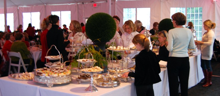 The image shows a catered event inside a large tent with people gathered around tables filled with various desserts and decorative greenery.