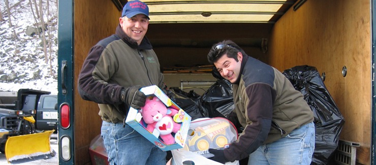 Two people are unloading toys from a vehicle's cargo area, smiling at the camera, with snow on the ground indicating winter season.