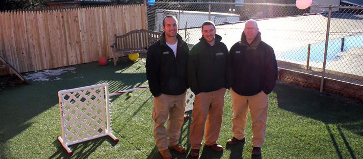 Three people are standing in an outdoor area with synthetic grass, smiling towards the camera, with a wooden fence and playground equipment in the background.