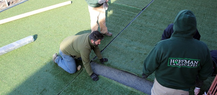 Three persons are installing artificial turf. Two are kneeling and adjusting the turf edges, while another stands holding turf. They wear work uniforms.