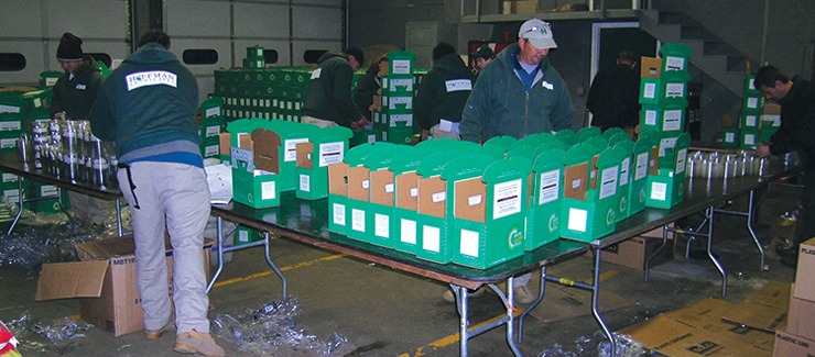 Several people are assembling packages in a warehouse. Green boxes with labels are arranged on tables with a person inspecting the contents.