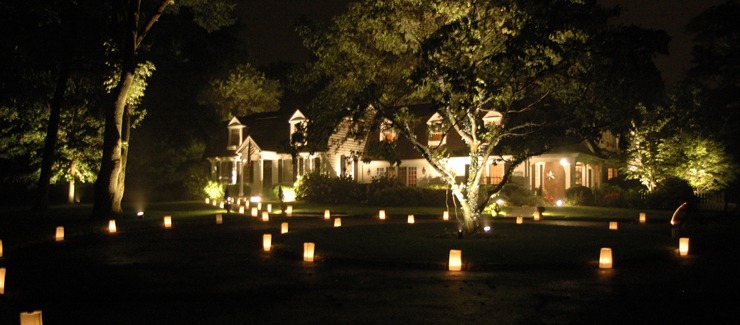 A large house is illuminated at night, with path lights and lit paper lanterns lining a driveway amidst trees, creating a warm and inviting atmosphere.