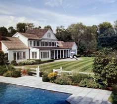 The image shows a large two-story house with white siding, shuttered windows, and multiple roofing sections, beside a manicured lawn and an outdoor swimming pool.