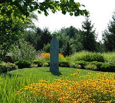 This image shows a vibrant garden with orange-yellow flowers in the foreground, a green sculpture in the center, and lush trees and shrubs in the background.
