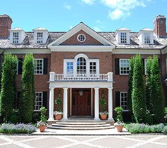 The image shows a large, elegant brick house with white trimming, a central portico, symmetrical windows, and manicured green hedges under a blue sky.