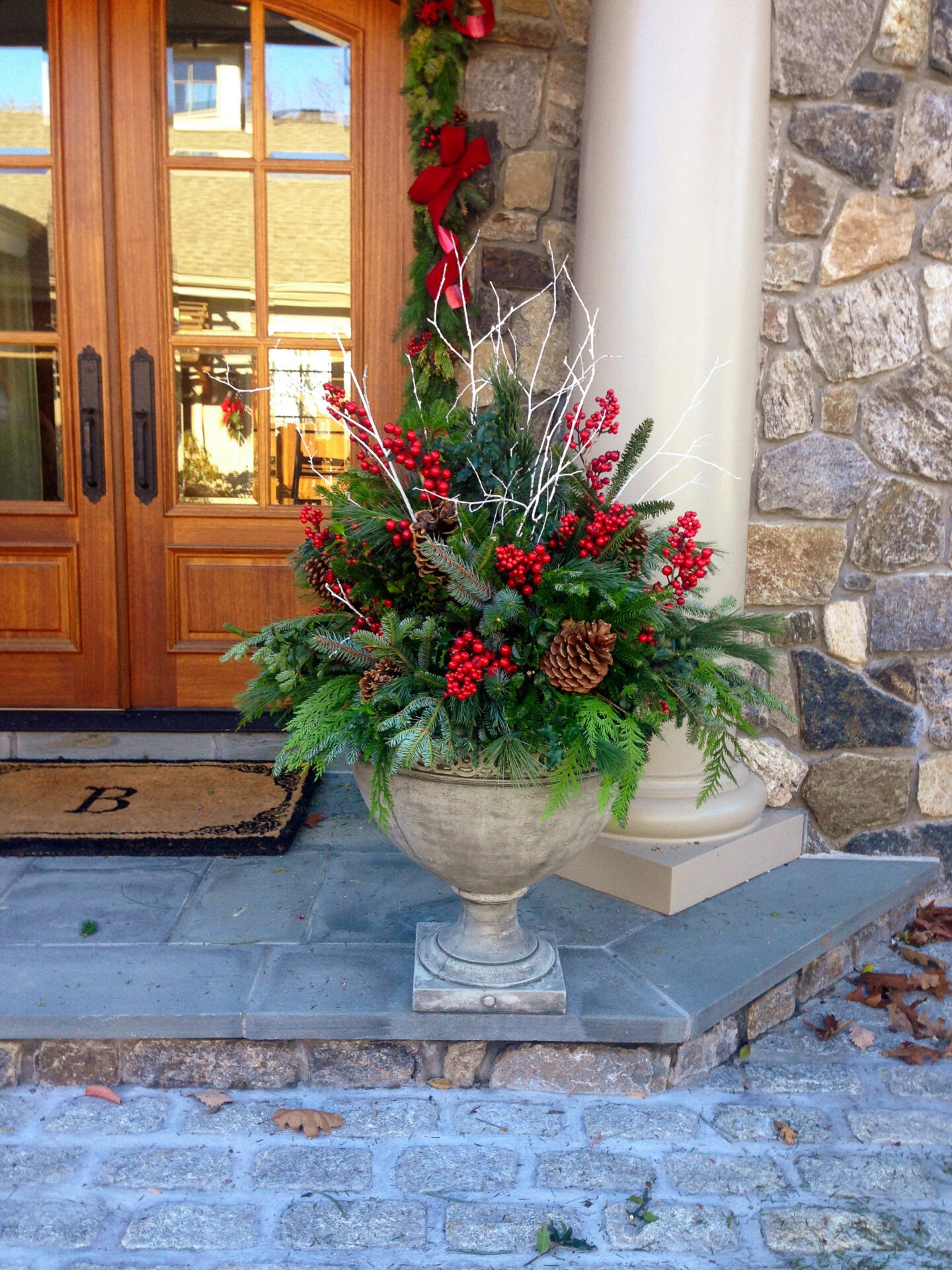 An ornate planter with evergreens, red berries, and pine cones sits on a porch, beside a wooden door adorned with a festive wreath.