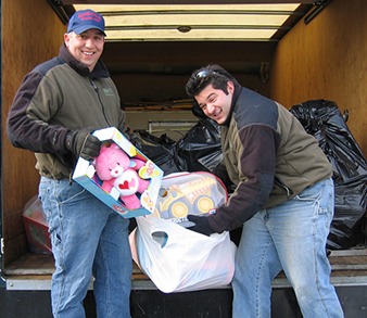 Two people are unloading donations from a truck, smiling at the camera. One holds toys, and black garbage bags are visible in the background.